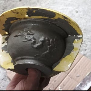 May include: A grey concrete planter, recently cast, held within a yellow mould. The planter has a rounded shape with a decorative relief design. The mould's edges show remnants of the casting process.