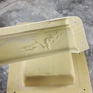 May include: A light yellow plaster moulding with a floral relief design. The moulding is rectangular with rounded corners and several small holes. The moulding is resting on a matching rectangular base.