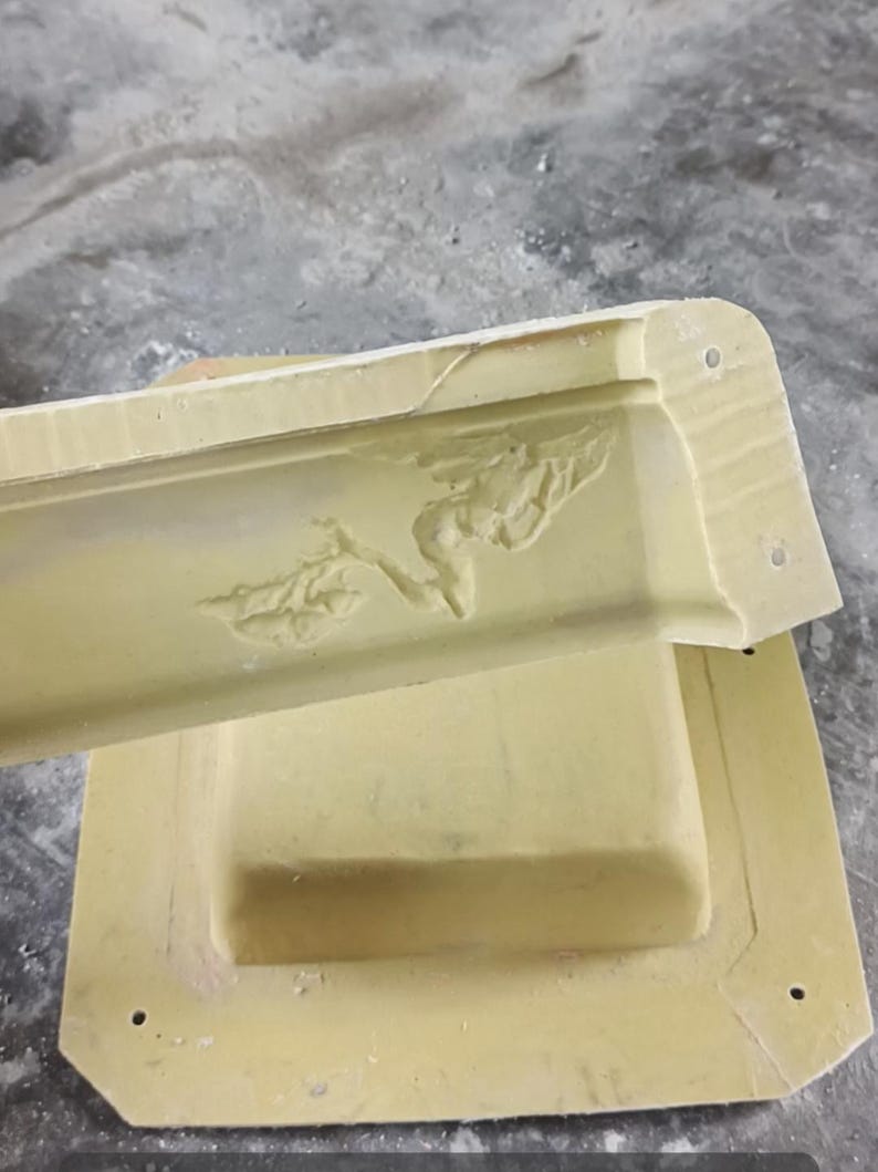 May include: A light yellow plaster moulding with a floral relief design. The moulding is rectangular with rounded corners and several small holes. The moulding is resting on a matching rectangular base.