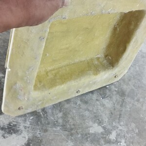 May include: A light yellow, angular object with a textured surface. The object has a rectangular shape with a recessed interior and is held by a hand. Small metal fasteners are visible along the edges.