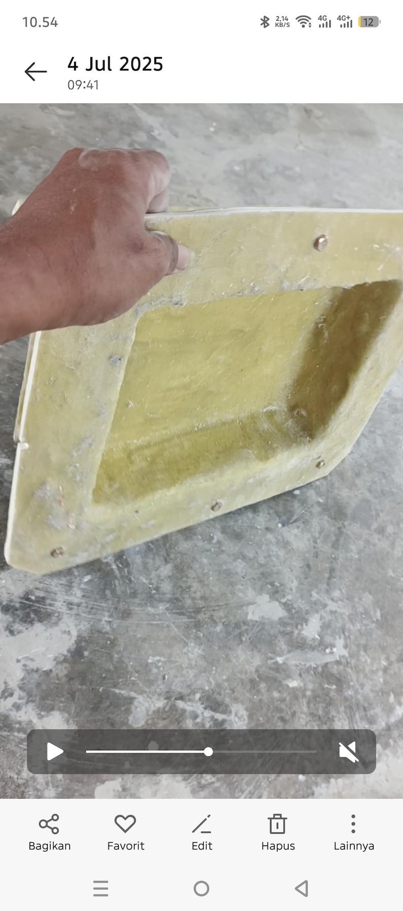 May include: A light yellow, angular object with a textured surface. The object has a rectangular shape with a recessed interior and is held by a hand. Small metal fasteners are visible along the edges.