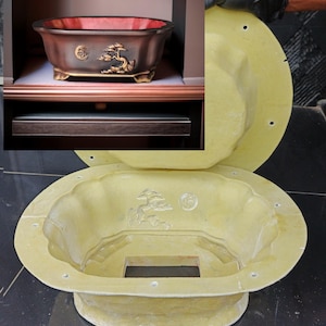 May include: A dark brown bonsai pot with gold detailing and a red interior. The pot, with a tree design, sits on a shelf. A mold of the pot is also visible, showing its shape and design.