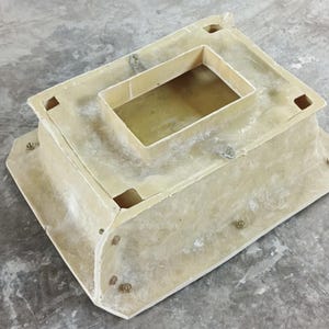 May include: A beige rectangular structure with a recessed top and a smaller rectangular opening. The structure has rounded corners and is secured with multiple bolts. The object is on a grey concrete surface.