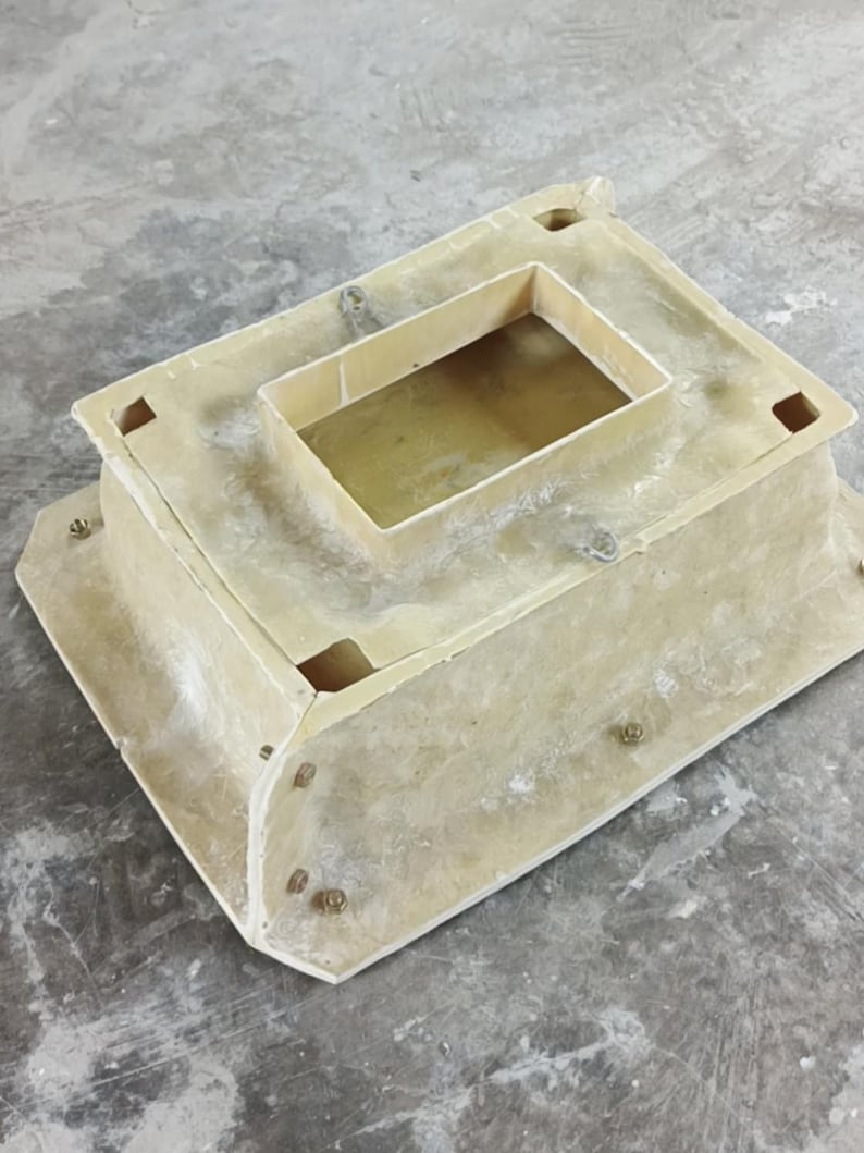 May include: A beige rectangular structure with a recessed top and a smaller rectangular opening. The structure has rounded corners and is secured with multiple bolts. The object is on a grey concrete surface.