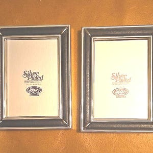 May include: Two rectangular silver-plated brass photo frames with black borders. Each frame has a cream-colored interior and the text "Silver Plated" and a logo. The frames are displayed on a brown surface.