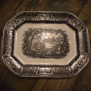 May include: An octagonal ceramic serving platter featuring a black and white transferware print. The center depicts a landscape with palm trees, a building, and an urn. The wide border is decorated with a detailed floral and scrollwork pattern.