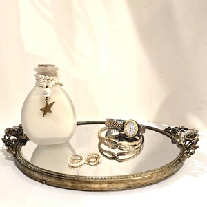 May include: A round, antique-style mirrored tray with ornate metal handles holds a frosted glass bottle adorned with a pearl necklace and a gold star charm, a two-tone metal wristwatch, a gold-toned bracelet, and a pair of small pearl hoop earrings.