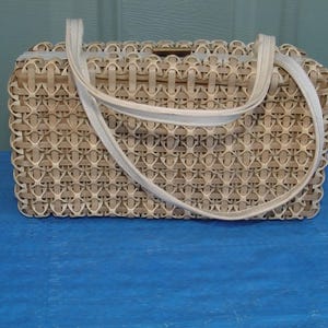 Op de afbeelding: Een vintage geweven handtas met een rechthoekige vorm en een crèmekleurige handgreep. De tas is gemaakt van lichtbruin geweven materiaal met een herhalend patroon. De handgreep is een lichtere crèmekleur.