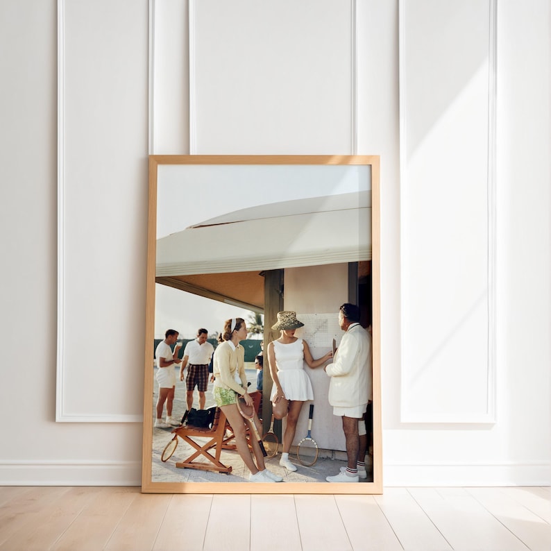 May include: A framed photograph depicts a vintage tennis scene. Several people in tennis attire are gathered near a structure. The image has a warm color palette, with a wooden frame and a white wall background.