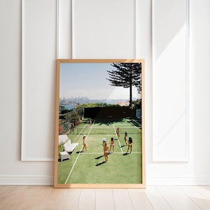 May include: A framed print featuring a tennis court scene with several people playing. The court is green with white lines, and the background shows a cityscape and blue sky. The frame is a light wood color.