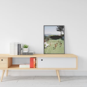 May include: A framed print depicts a tennis court scene with several people playing. The court is green with white lines, and the players are in the middle of a match. The background shows a city skyline and a large tree. The frame is black.