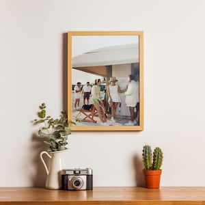 May include: A framed photograph of a group of people in white clothing, standing near a building. A vintage camera, a white pitcher with greenery, and a small cactus in an orange pot sit on a wooden surface.