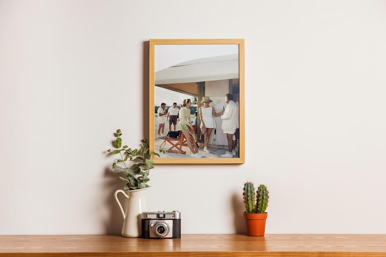 May include: A framed photograph of a group of people in white clothing, standing near a building. A vintage camera, a white pitcher with greenery, and a small cactus in an orange pot sit on a wooden surface.