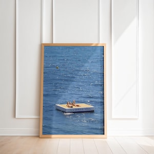 May include: A framed print showing a rectangular floating platform on blue water. Two figures are sunbathing on the platform. The platform has the text "LA DOLCE VITA" printed on it. The frame is a light brown colour.