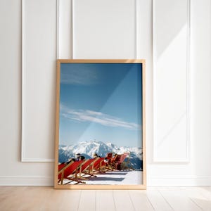 May include: A framed print featuring a scenic mountain landscape with snow-covered peaks under a bright blue sky. Several red deck chairs are arranged on a snowy slope, suggesting a ski resort or mountain retreat.