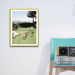May include: A framed print with a yellow border depicts a tennis court scene with players. A yellow radio sits on a stack of magazines next to a teal chair. A white lamp is also in the image.