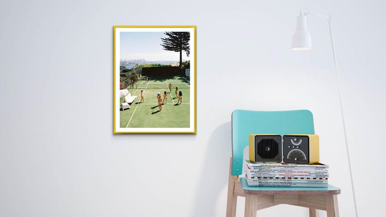 May include: A framed print with a yellow border depicts a tennis court scene with players. A yellow radio sits on a stack of magazines next to a teal chair. A white lamp is also in the image.