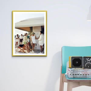 May include: A framed photograph with a yellow border depicts a group of people in white and light green clothing. A vintage-style radio sits on a stack of magazines next to a teal chair. A white lamp is positioned above.