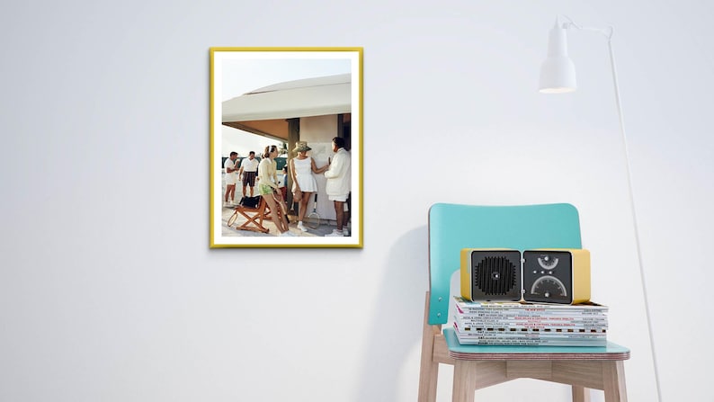 May include: A framed photograph with a yellow border depicts a group of people in white and light green clothing. A vintage-style radio sits on a stack of magazines next to a teal chair. A white lamp is positioned above.