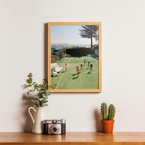 May include: A framed print of a tennis court scene with players, set against a city backdrop. The print is in a light wood frame, displayed on a wooden surface with a vintage camera, a white pitcher with greenery, and a potted cactus.