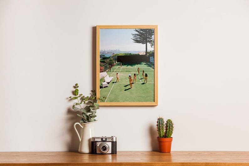 May include: A framed print of a tennis court scene with players, set against a city backdrop. The print is in a light wood frame, displayed on a wooden surface with a vintage camera, a white pitcher with greenery, and a potted cactus.