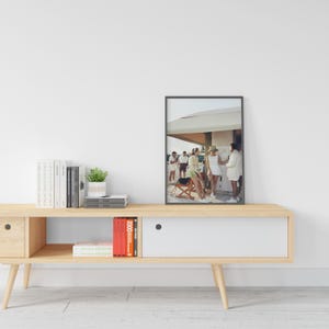 May include: A framed photograph featuring a group of people in white and light-colored clothing, standing near a building with a white awning. The photograph is displayed on a light wood console table, alongside books and a potted plant.