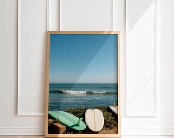 May include: A framed print featuring a coastal scene with a clear blue sky and ocean waves. Two surfboards, one mint green and one white, rest on the shore. The print is in a wooden frame, leaning against a white wall.
