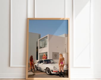 May include: A framed print showing a white sports car with black racing stripes, parked in front of a modern white building. Three people in swimwear are near the car. The sky is blue.