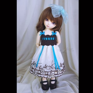 May include: A small doll wearing a white dress with brown crocheted bodice and blue ribbon accents. The doll has brown hair and blue eyes and is wearing a blue bow headband.