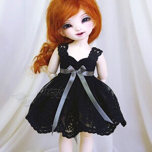 May include: A doll with red hair wearing a black lace dress with a gray satin ribbon. The doll is wearing black shoes with a lace trim.