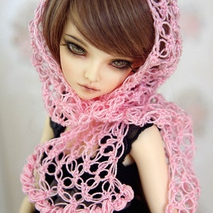 Puede incluir: Una mu&ntilde;eca con un chal de ganchillo rosa y un vestido negro. El chal tiene un delicado patr&oacute;n floral y un borde festoneado. La mu&ntilde;eca tiene el pelo casta&ntilde;o y los ojos azules.