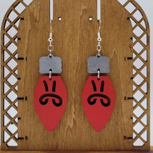 May include: A pair of red and gray wooden earrings shaped like Christmas light bulbs. Each earring has a gray rectangular top and a red teardrop-shaped bottom with a black face design. The earrings hang from silver-colored hooks.