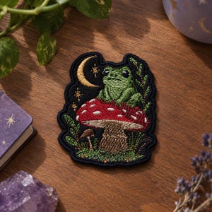 Puede incluir: Un parche bordado que muestra una rana verde sentada sobre una seta roja grande con puntos blancos. El diseño incluye una luna creciente dorada, pequeñas estrellas y follaje verde sobre un fondo negro con un borde cosido oscuro.