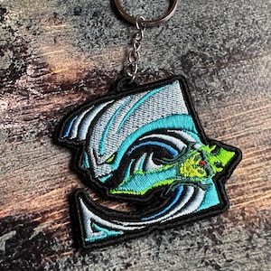 Op de afbeelding: Geborduurde sleutelhanger met een gestileerd drakenontwerp in blauw, groen en wit, omlijnd in zwart. De draak heeft rode ogen en is bevestigd aan een zilveren sleutelhanger.