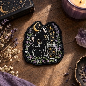 May include: An embroidered patch featuring a black cat sitting next to a glass jar filled with glowing yellow stars. The design includes a golden crescent moon above the cat, surrounded by purple and green floral accents on a black background.