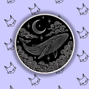 Puede incluir: Un parche bordado en blanco y negro con una ballena bajo una luna creciente y estrellas. El diseño está rodeado de nubes y olas. El fondo es de color lila claro con ilustraciones repetidas de caras de gato.