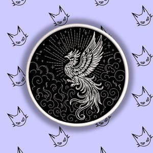 May include: A circular black and white embroidered patch featuring a phoenix rising from stylized clouds. The phoenix is detailed with white thread, set against a black background. The patch has a white border and is set against a light purple background with cat head doodles.