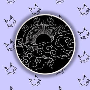 Könnte beinhalten: Ein schwarz-weißes kreisförmiges Design mit Sonne, Wolken und einem Halbmond. Das Design ist von einem weißen Rand umgeben und vor einem hellvioletten Hintergrund mit sich wiederholenden Katzenkopf-Illustrationen platziert.
