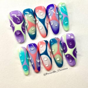 May include: A set of ten long, oval-shaped artificial nails. Each nail features a unique design with a base color of purple, blue, pink, or green, overlaid with abstract patterns and clear, droplet-shaped embellishments. The text "@PressedBy_Christina" is visible.
