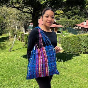 Puede incluir: Un bolso tote tejido azul con un diseño de rayas coloridas. El bolso tiene correas largas y trenzadas en tonos de azul, morado y rosa. El bolso se sostiene frente a un fondo verde y herboso con árboles y casas.