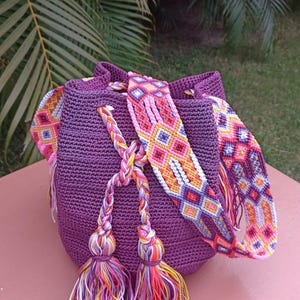Puede incluir: Bolso de crochet morado hecho a mano con una correa estampada y borlas coloridas. La correa presenta un diseño geométrico en rosa, naranja, amarillo y blanco. El bolso tiene un cierre de cordón y las borlas son una mezcla de rosa, amarillo y morado.
