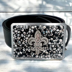 Puede incluir: Cinturón de cuero negro con una hebilla rectangular plateada. La hebilla está incrustada con pequeños cristales brillantes y presenta un diseño de flor de lis en el centro. La flor de lis tiene una gema rosa en el centro.