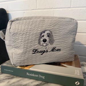 May include: A gray and white striped cosmetic bag with an embroidered dog face and the text "Doug's Mom." The bag is resting on a book titled "Resident Dog."