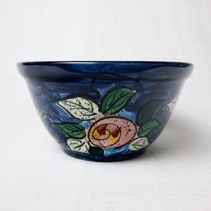 May include: A dark blue ceramic bowl with a floral design. The bowl features a pink and yellow rose surrounded by green and white leaves. The bowl is a decorative piece.