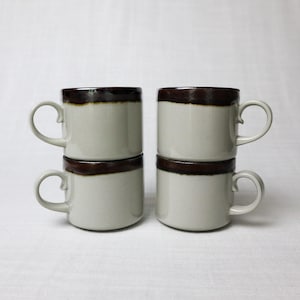 May include: Four stacked ceramic mugs. Each mug is off-white with a dark brown band around the top. The mugs have a simple, curved handle. The mugs are arranged in two stacks of two, set against a white background.
