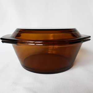 May include: A vintage amber-colored glass casserole dish with a matching lid. The dish has a rounded base and a flared rim with handles. The lid sits flush on top, creating a cohesive design. This kitchenware is ideal for baking and serving.