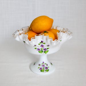 May include: A white, ruffled-edge pedestal bowl filled with bright yellow lemons. The bowl is decorated with hand-painted purple flowers and green leaves, and has a gold trim.