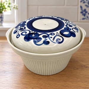 May include: A cream-colored ceramic bowl with a matching lid featuring a dark blue hand-painted floral pattern. The bowl has a ribbed exterior texture, while the lid displays a central blue circle surrounded by intricate swirling blue motifs.