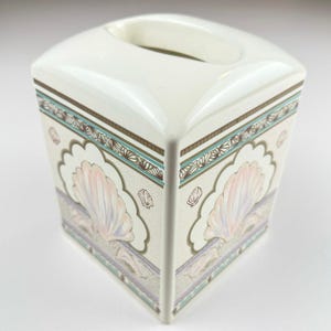 May include: A square, off-white ceramic tissue box with a decorative seashell design. The box features a scalloped border with pink, purple, and blue seashell motifs. Brown and teal accents frame the seashell pattern.