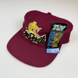 Peut inclure: Casquette de baseball bordeaux avec une visière plate. Le chapeau présente un texte brodé en or et un logo "Sun Devils". Une étiquette sur le côté indique "Top of the World".